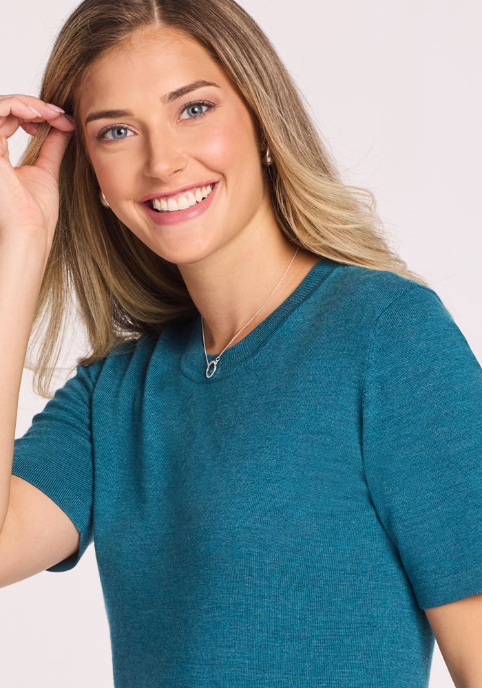 A smiling woman with long, light brown hair wears the Woolx Anna Crew Sweater in Harbor Teal and a silver necklace. She poses with a hand near her face against a plain, light background.
