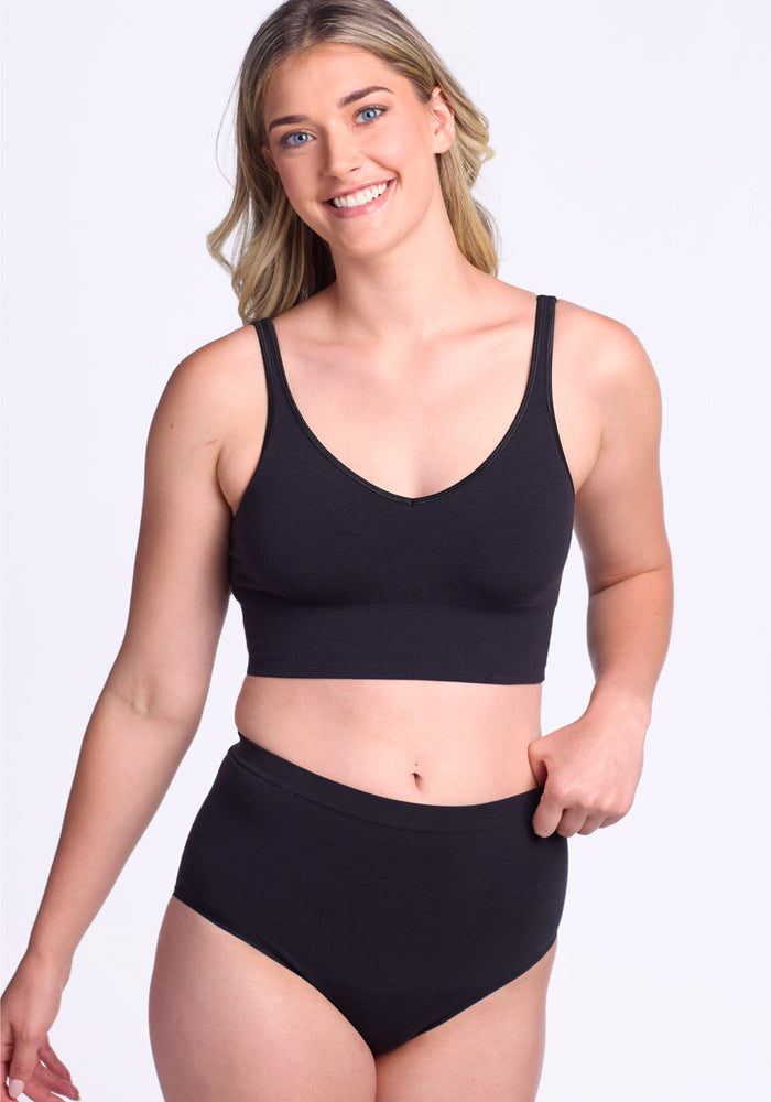 A smiling woman with long blonde hair models the Woolx Mabel Full Coverage Underwear in black, featuring a matching sleeveless bralette, against a plain white background, highlighting seamless comfort and style.