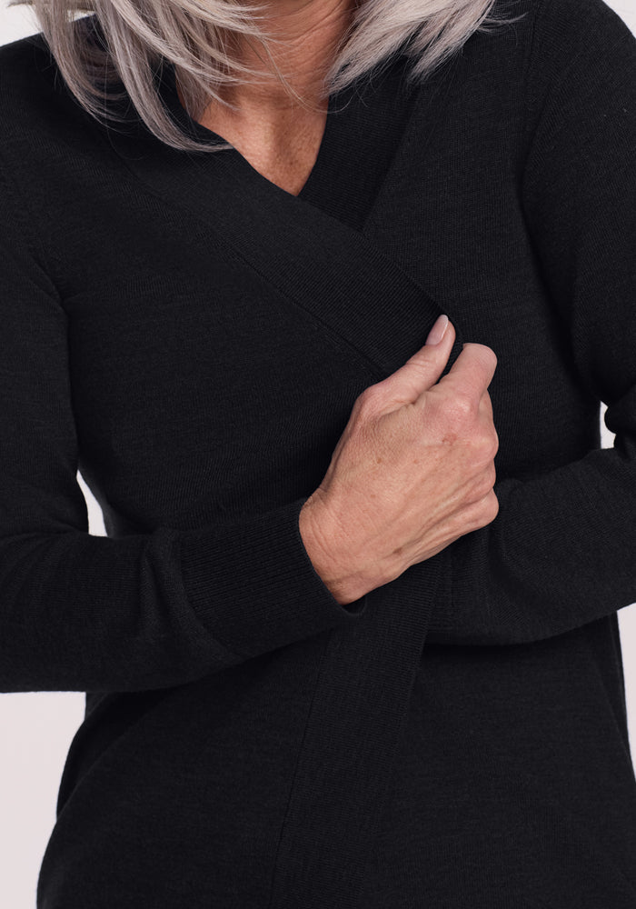 A person with light-colored hair wears the Woolx Macie Sweater in black, holding the open front closed at their chest with their right hand. The face is mostly out of frame.