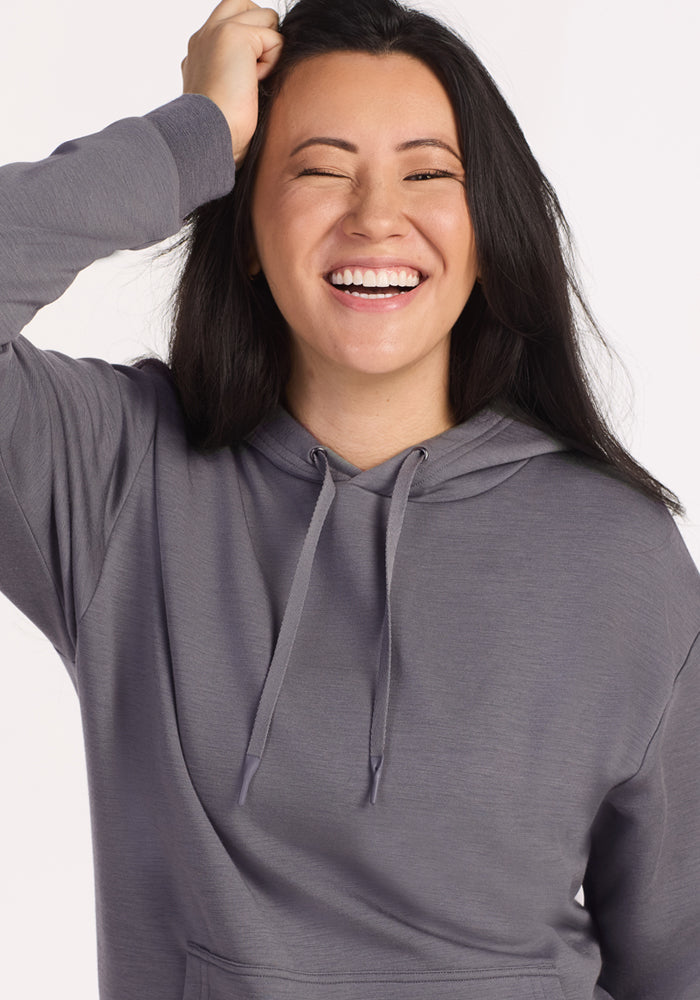 A woman with long dark hair in the Woolx Avery Hoodie - Arctic Dusk smiles brightly and winks, playfully touching her head against a light background, radiating the cozy, year-round comfort of Merino wool.