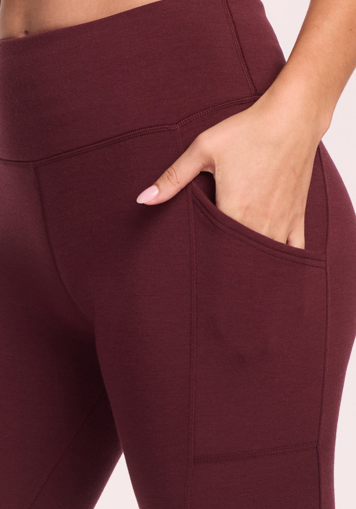 A close-up shows a person wearing Woolx Piper Pocket Leggings in Toasted Garnet, featuring a high waist and side pocket with their hand partially tucked in—ideal as a women's Merino wool baselayer for cold weather.