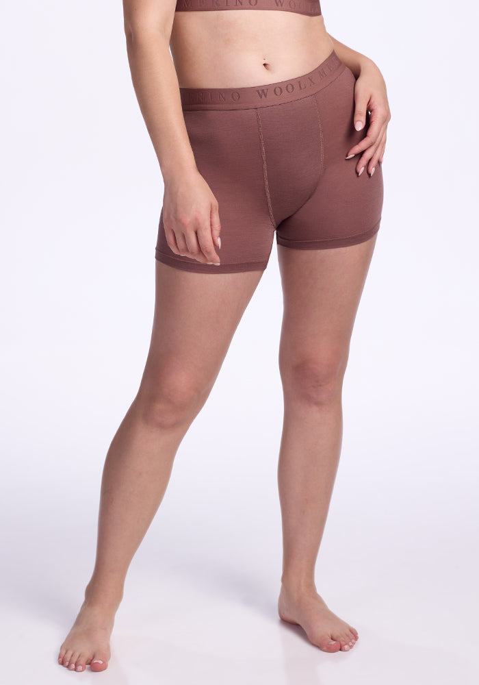 A person stands barefoot against a plain white background, wearing the Woolx Elliott Boyshort in Nutmeg and a matching brown sports bra, with one hand on their hip and the other by their side. 
