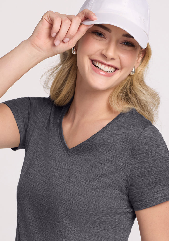 A smiling woman in the Woolx Junie V Neck - Cinder Melange, a relaxed gray Merino wool tee, wears a white cap and touches its brim. She has light hair and looks at the camera against a plain background.