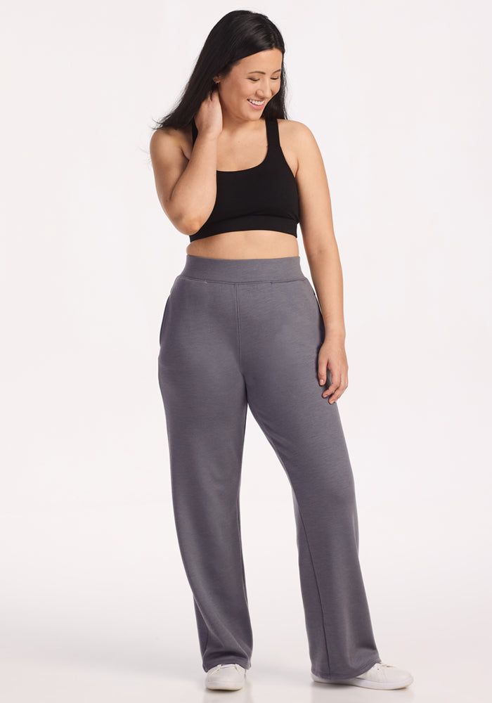 A woman with long dark hair wears a black crop top, white sneakers, and Woolx Bree Lounge Pants in Arctic Dusk. Smiling and touching her hair, she stands against a plain white background.