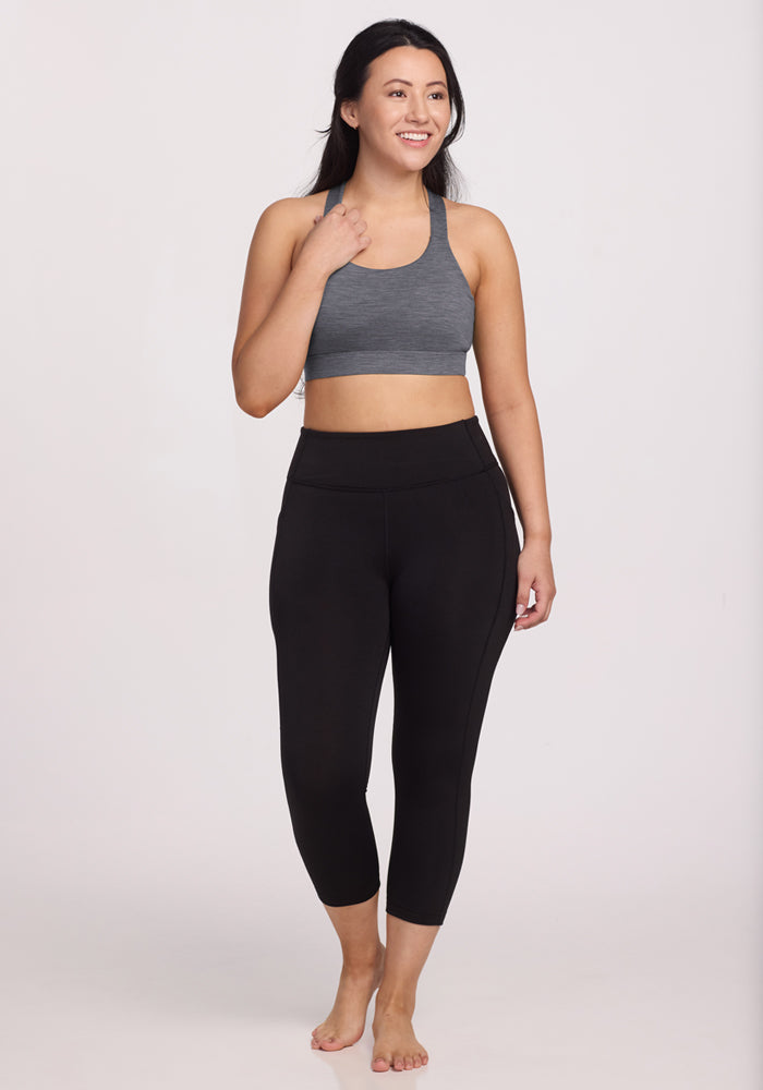 A woman stands barefoot against a plain background, smiling as she holds her hair with one hand. She wears the Woolx Livi Bra in Graphite Heather and black high-waisted athletic leggings for all-day comfort.