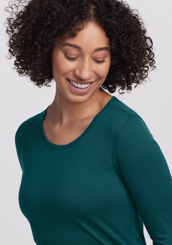 A woman with curly dark hair, wearing the Woolx Remi Long Sleeve T-Shirt in Riverbank Green, smiles while looking down against a plain light gray background.