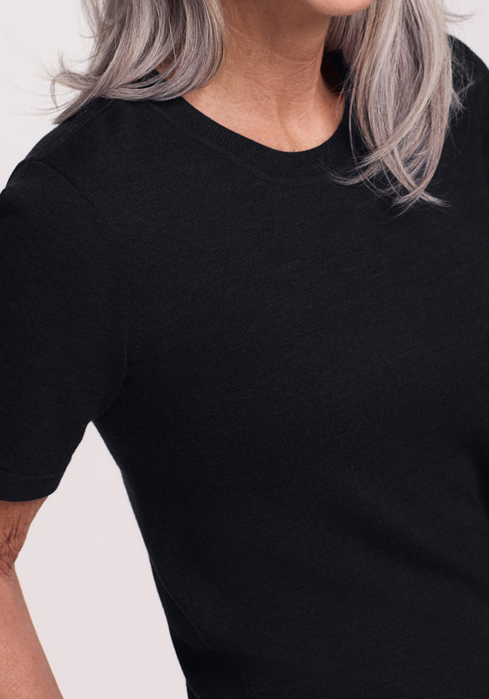 A close-up shows a person with long gray hair, partially out of frame, wearing the Woolx Anna Crew Sweater in black from shoulders to waist.