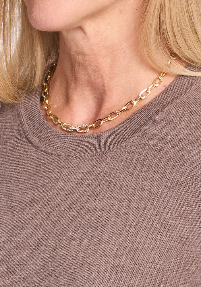 Close-up of a person wearing a gold chain necklace with an oval textured clasp, paired with the Woolx Anna Crew Sweater in Simply Taupe. The person's light blonde hair is partially visible.