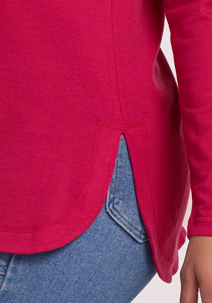 A close-up of a person in the Woolx Hazel Tunic - Poppy Spark, a bright pink merino wool top with a curved hem and side notch, paired with blue denim jeans. Focus is on the temperature-regulating fabric and hip hem detail.