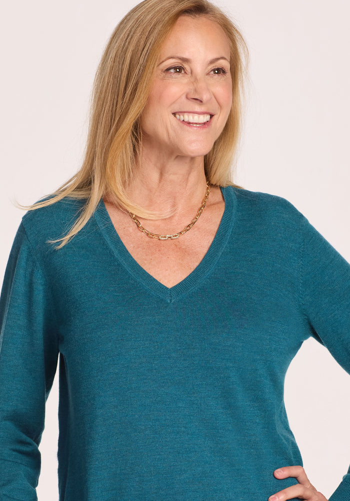 A smiling woman with long blonde hair wears the Woolx Francesca Sweater in Harbor Teal and a gold chain necklace, standing against a plain light background.