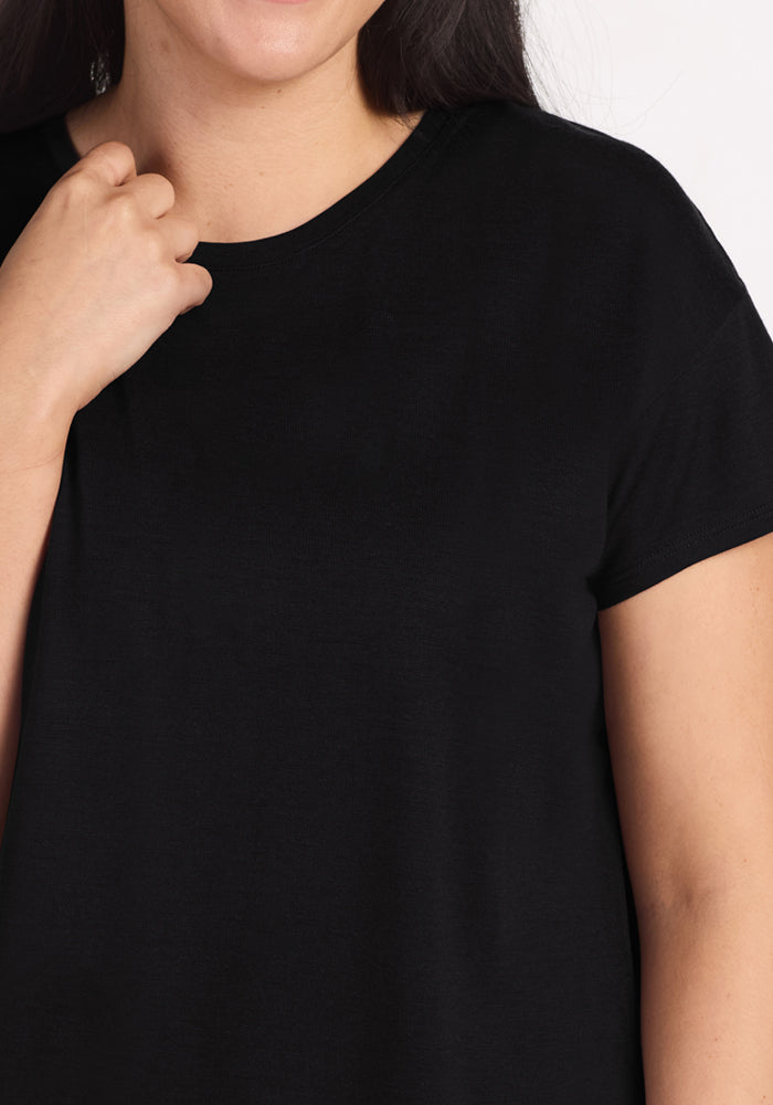 A woman wears the Woolx Billie Lounge Tee in black, shown from shoulders to upper torso with one hand near her collarbone and a partial smile.