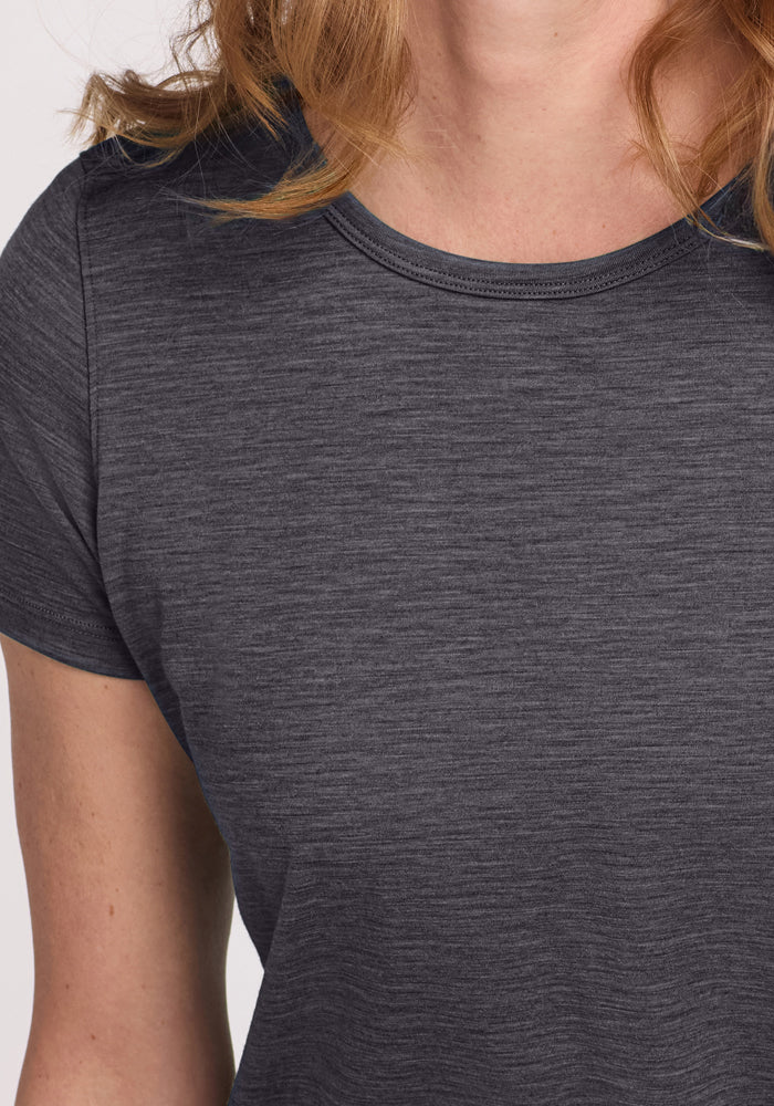 A close-up shows a person in the Woolx Liza Crew Neck Tee - Cinder Melange, a dark gray, short-sleeved Merino wool shirt. The lightweight performance tee is worn with light brown hair partially visible and the face mostly out of frame.