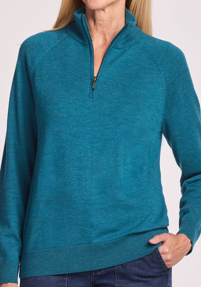 A woman wears the Woolx Collins Half Zip Sweater in Harbor Teal with her left hand in her dark blue jeans pocket, shown from the chin down.