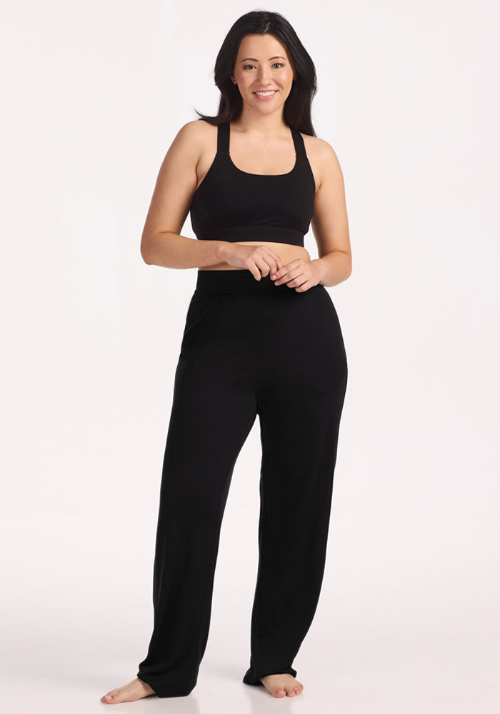 A woman with long dark hair stands barefoot against a white background, smiling. She is wearing a black crop top and Woolx Maya Lounge Pants - Black, designed for comfort and style with moisture-wicking fabric.