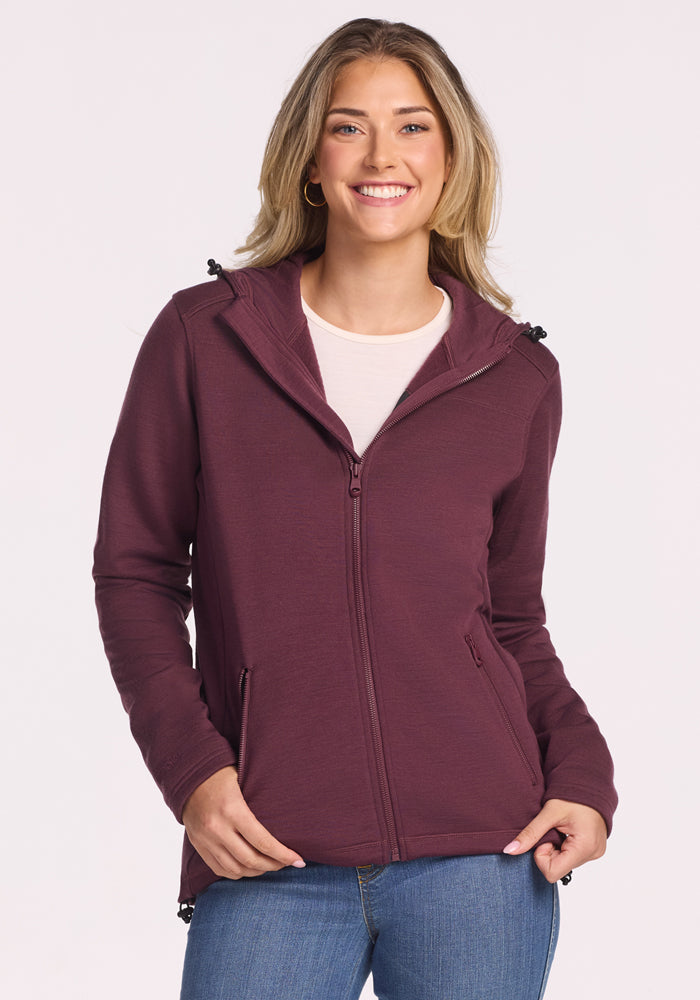 A smiling woman with long blonde hair wears the Woolx Cubby Hooded Sweatshirt in Grape Harvest over a light top and blue jeans, standing against a plain light background. 