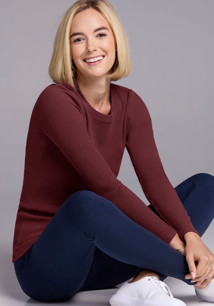 A smiling blonde woman sits cross-legged on the floor, wearing the Woolx Hadley Ribbed Crew in Toasted Garnet, navy leggings, and white sneakers, against a plain gray background—perfect for lightweight layering and travel.
