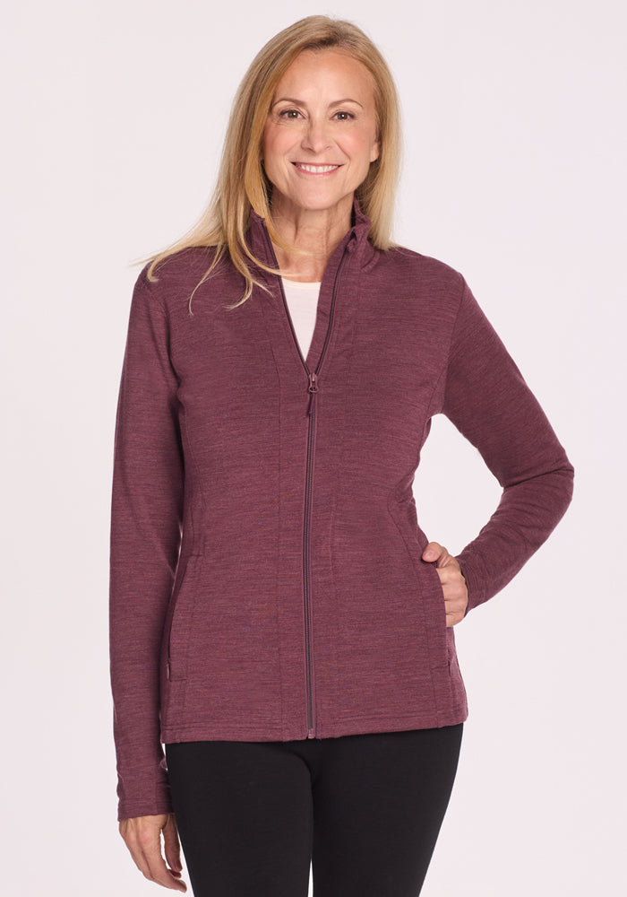 A woman with long blonde hair smiles with one hand on her hip, wearing the Woolx Finley Zip Up Sweatshirt in Grape Harvest Melange and black pants, standing against a plain light background. 
