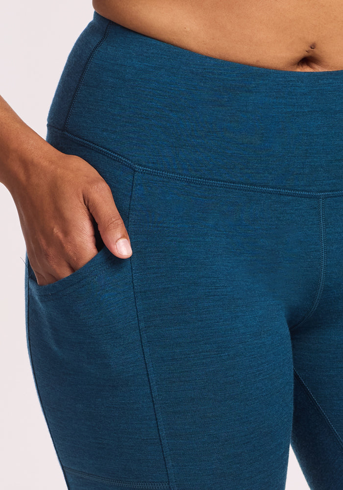 A person wears Woolx Piper Pocket Leggings in Majolica Blue, with their right hand in the side pocket. Only the torso and upper legs are shown.