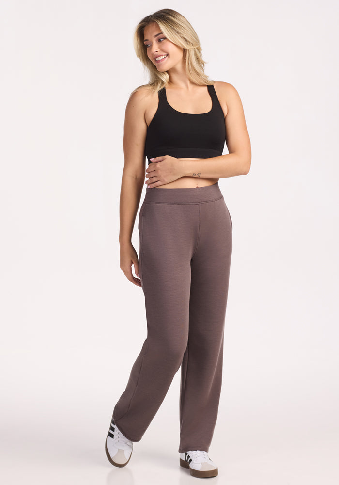 A woman with blonde hair wears a black tank top, white sneakers, and Woolx Bree Lounge Pants in Plum Truffle. She stands against a white background, smiling and looking to her left with one hand relaxed by her side.