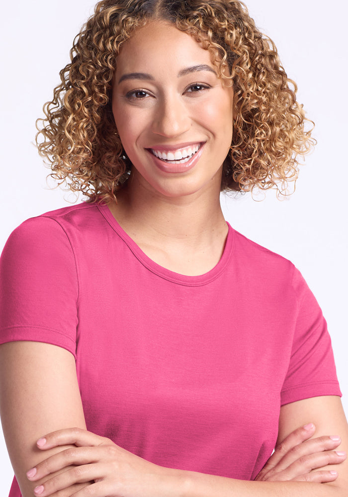 A smiling woman with curly, shoulder-length hair wears the Woolx Liza Crew Neck Tee in Raspberry Fizz, standing against a light background with her arms crossed.