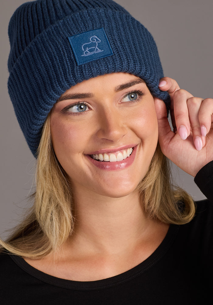 A smiling woman wears the Woolx Ria Chunky Beanie in Starry Night, featuring an embroidered animal logo, with a black top, holding her winter accessory against a plain neutral background.