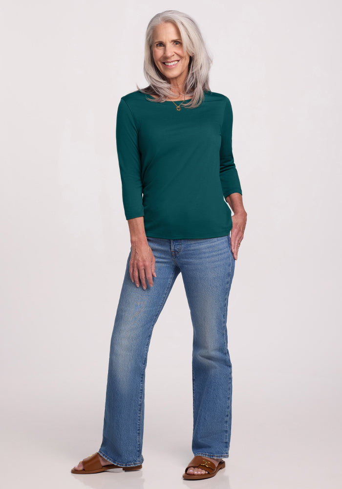 A smiling older woman with long gray hair stands against a light background, wearing the Woolx Jenny 3/4 Sleeve Crew Neck in Riverbank Green, blue jeans, and brown sandals. One hand rests on her hip while the other is by her side.