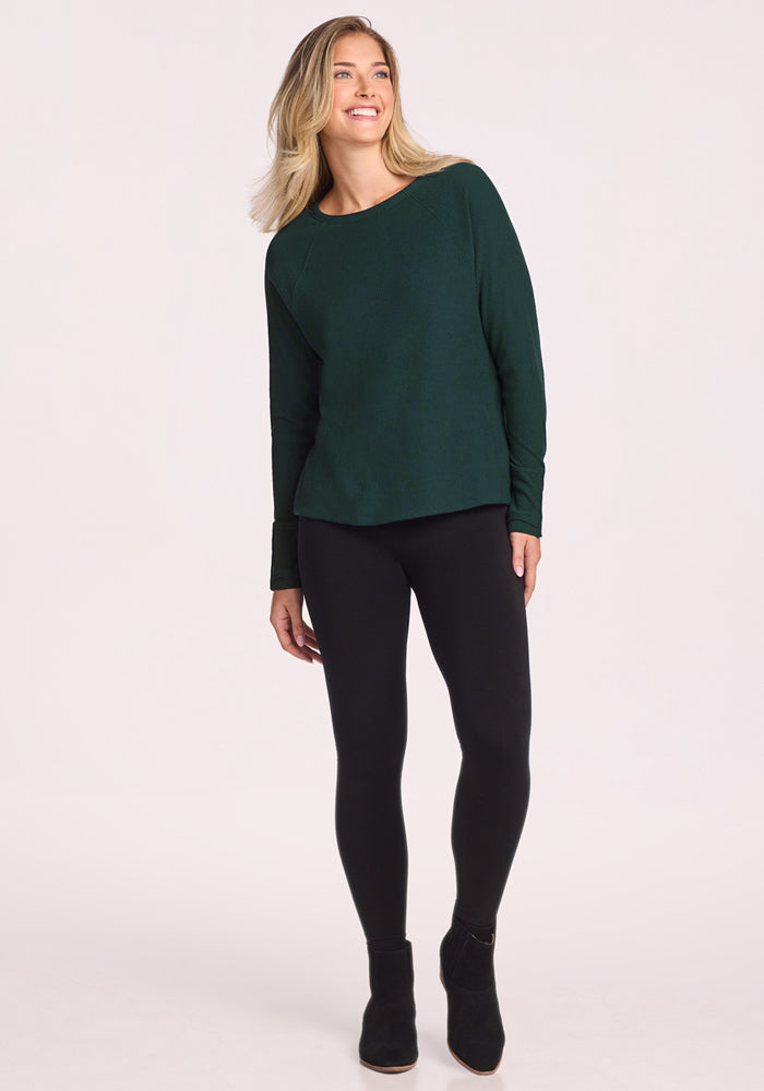 A woman stands smiling, wearing the Woolx Ainsley Sweater in Deep Timber, paired with black leggings and black ankle boots against a plain light background.