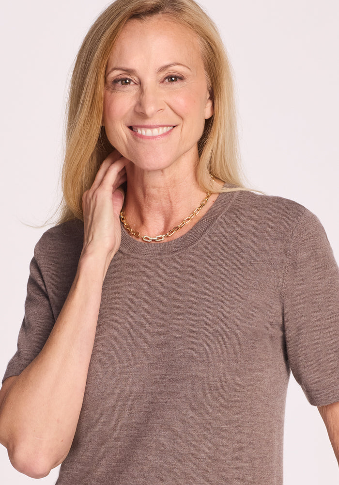 A smiling woman with long blonde hair wears the Woolx Anna Crew Sweater in Simply Taupe and a gold chain necklace, standing against a light background with one hand touching her neck.