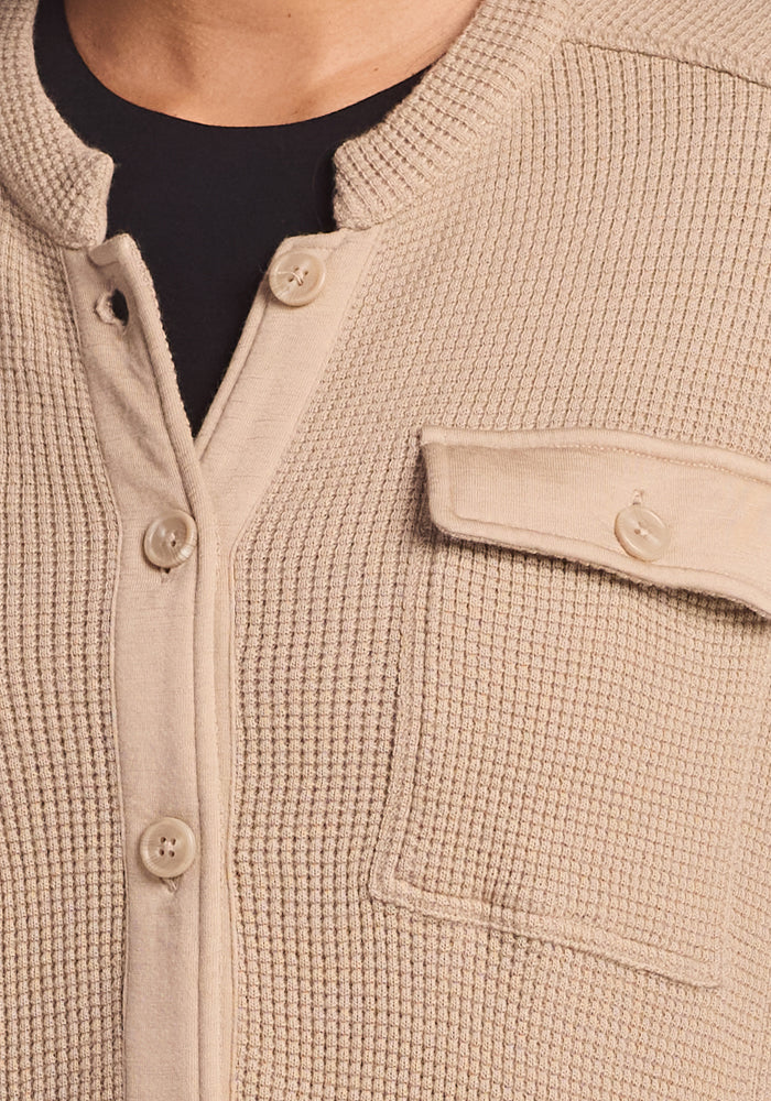 The Woolx Oaklynn Waffle Shacket in Biscotti features a beige, textured waffle knit with a button-up front and chest pocket flap. Layered over a black top, it offers a cozy, casual look while revealing part of the neck.