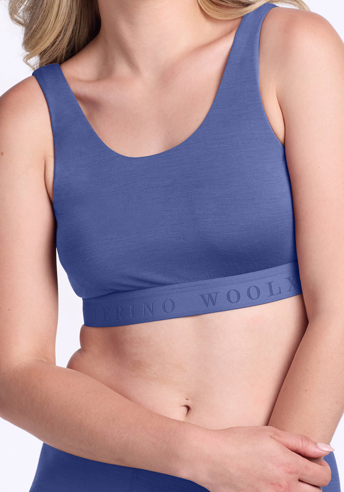 A woman wears the Woolx Aubrey Lounge Bra in Cloudy Bay, a sleeveless light blue crop top with an embossed Woolx logo band. Her arms are relaxed and her face is partially out of frame.