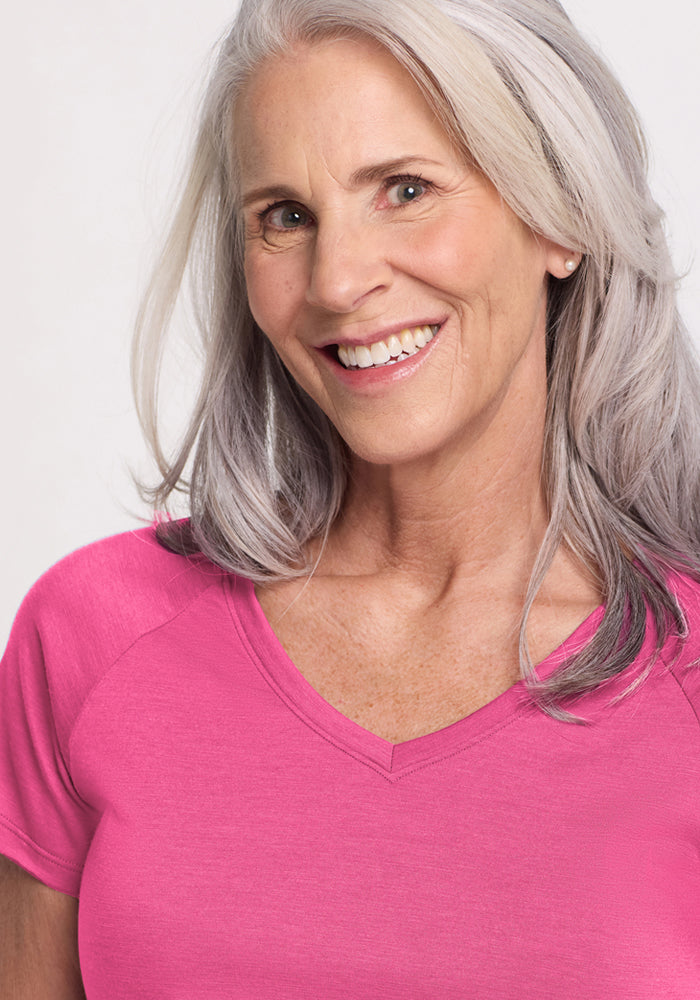 A smiling older woman with long gray hair wears the Woolx Mia V Neck - Raspberry Fizz, a bright moisture-wicking Merino wool t-shirt, standing against a plain light background.