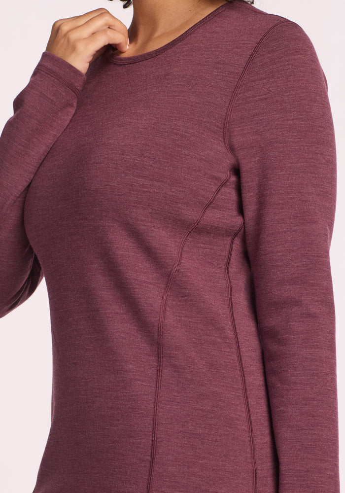 A person wearing the Woolx Elsa Long Sleeve in Grape Harvest Melange, a merino wool base layer with long sleeves and visible seams, is shown from shoulders to waist with one hand near their neck.