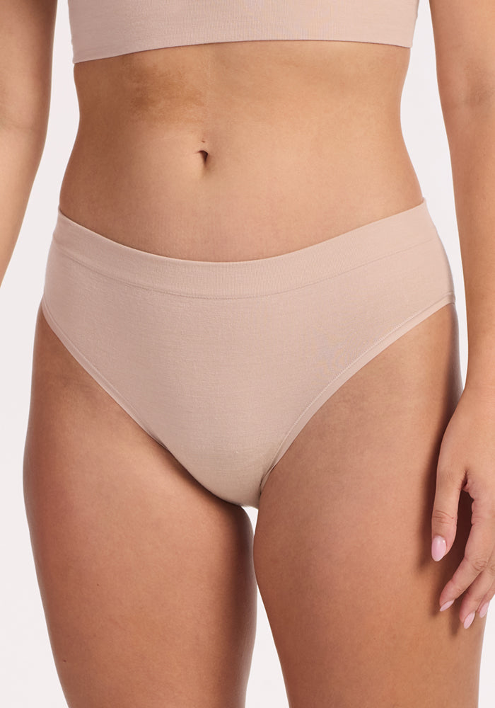 Model wears the Woolx Maeve Bikini in Sand, a high-waisted seamless style, paired with a matching crop top. Shown from mid-torso to upper thighs against a plain background.