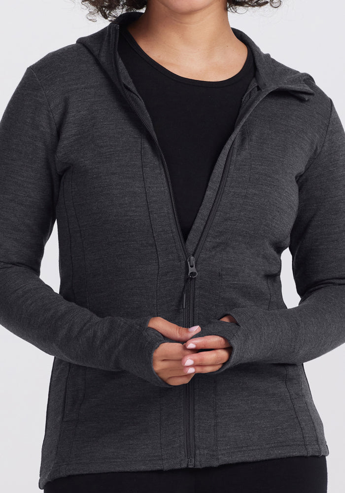 A person wears a Woolx Zoey Zip Up Hoodie in Cinder Melange over a black shirt, standing against a plain light background. The women's slim-fit hoodie is partially unzipped, with hands visible near the zipper and thumbholes showing.
