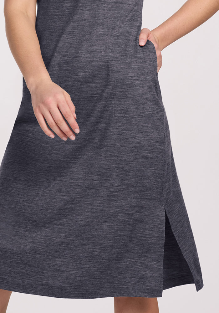 A person stands against a plain, light background in the Woolx Cassie Dress - Cinder Melange, a heather gray, knee-length Merino wool dress with a side slit and pocket. This travel-friendly piece resists wrinkles for comfort and style.