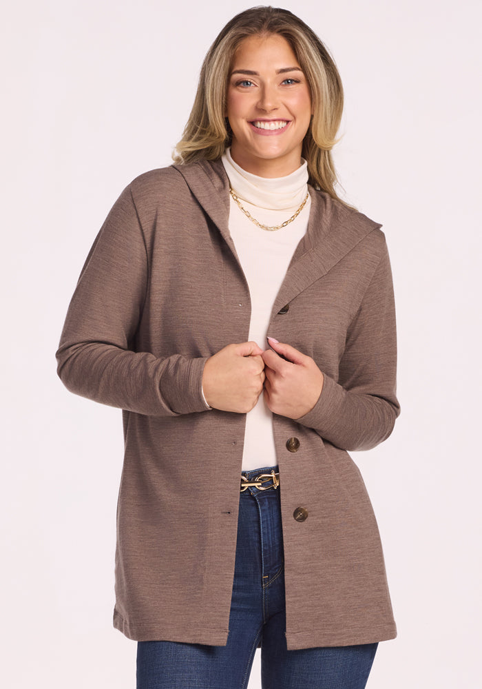A woman with long blonde hair smiles in the Woolx Edna Hooded Cardigan in Simply Taupe, styled over a white turtleneck, blue jeans, and a gold chain necklace—a cozy cold-weather look against a plain light background. 