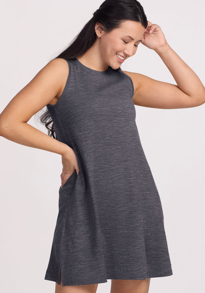 A woman with long dark hair smiles as she looks down, wearing the Woolx Clara Dress in Cinder Melange. The sleeveless, knee-length Merino wool dress stands out against a light background, highlighting its stylish and breathable summer design.