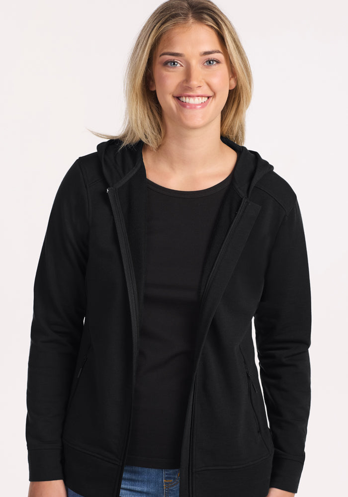 A young woman with long blonde hair smiles at the camera, wearing the Woolx Darby Zip Up Hoodie in black over a black shirt and blue jeans, standing against a plain light background.