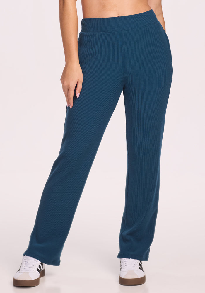 A person wears Woolx’s Luca Ribbed Pants in Majolica Blue with white sneakers featuring black stripes, standing against a plain light background. Only the lower half, showcasing the stylish wide leg pants, is visible.