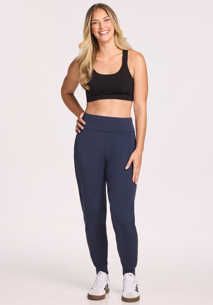 A woman with long blonde hair wears a black crop tank top, Woolx Willa Joggers in Deep Navy, and white sneakers. She is smiling and standing against a plain light background.