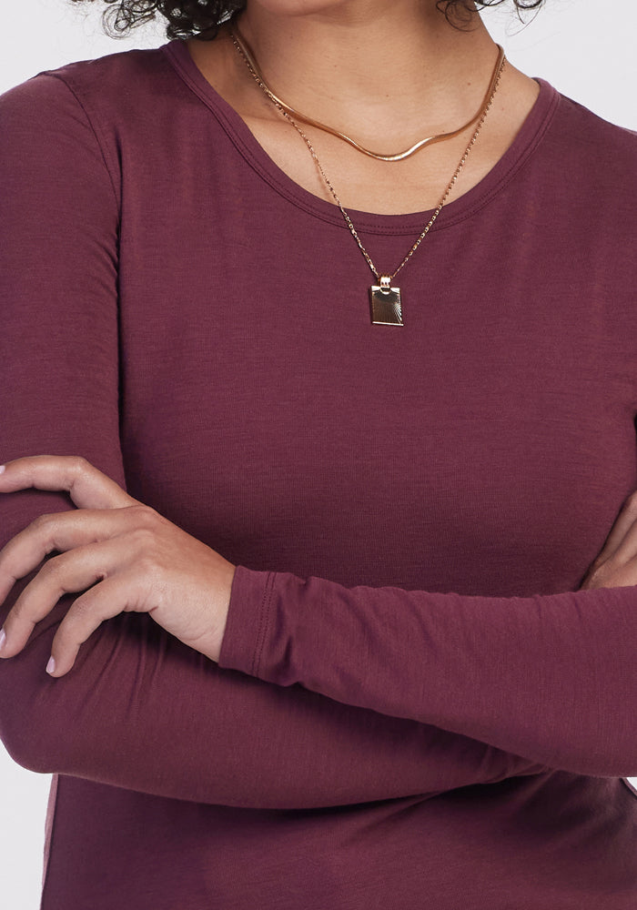A person with crossed arms wears the Woolx Remi Long Sleeve T Shirt in Grape Harvest, along with a gold chain and pendant necklace. The image is cropped to display just the upper torso and lower face.