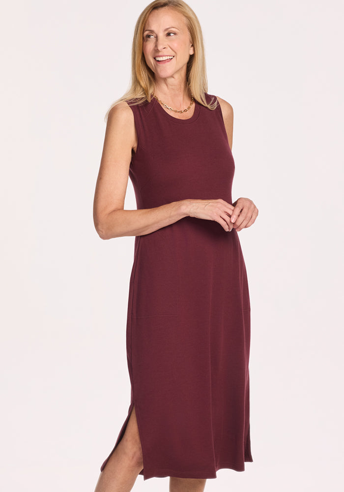 A woman with light skin and straight, shoulder-length blonde hair smiles in the Woolx Cassie Dress in Toasted Garnet—a sleeveless, knee-length Merino wool tank dress with side slits that's perfect for travel thanks to its temperature-regulating fabric.