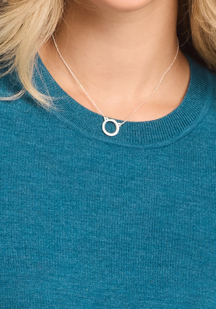 A person wears the Woolx Anna Crew Sweater in Harbor Teal and a delicate silver necklace with a small round pendant. The image is cropped to show only the lower face, neck, and upper chest.