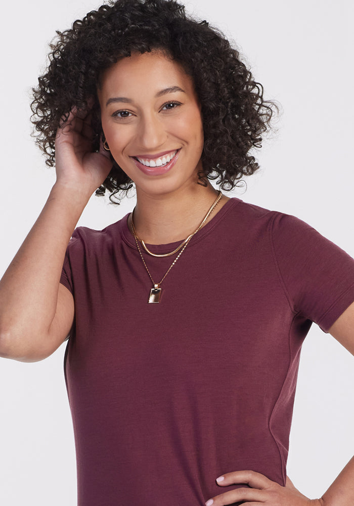 A woman with curly hair smiles, touching her hair. She wears the Woolx Liza Crew Neck Tee in Grape Harvest and layered gold necklaces, standing against a plain light background.