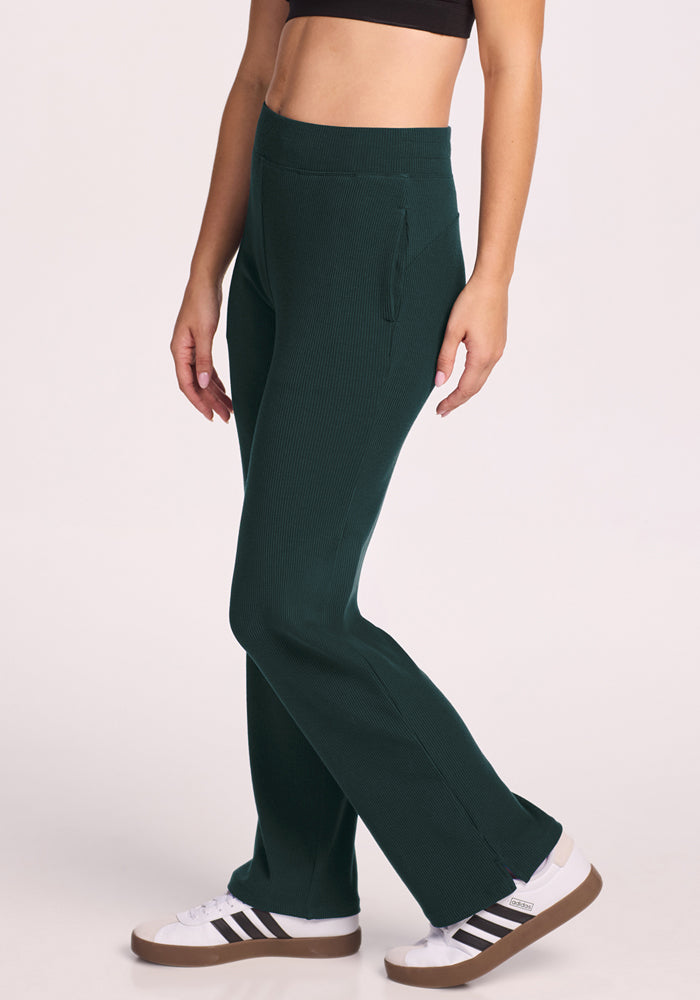 A person wearing Woolx Luca Ribbed Pants in Deep Timber, a black crop top, and white sneakers with black stripes stands against a light background. The person's face is not visible.