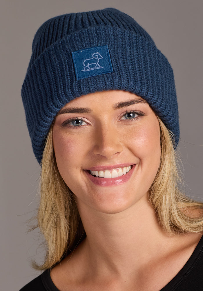 A smiling blonde woman wears the Woolx Ria Chunky Beanie in Starry Night—a dark blue knit hat with a rectangular animal logo patch. She pairs it with a black top, set against a neutral gray background.