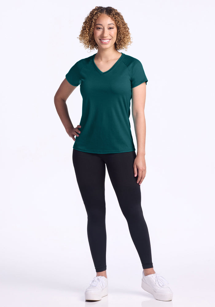 A smiling woman with curly hair wears the Woolx Mia V Neck in Riverbank Green, paired with black leggings and white sneakers. She stands with one hand on her hip against a plain white background.