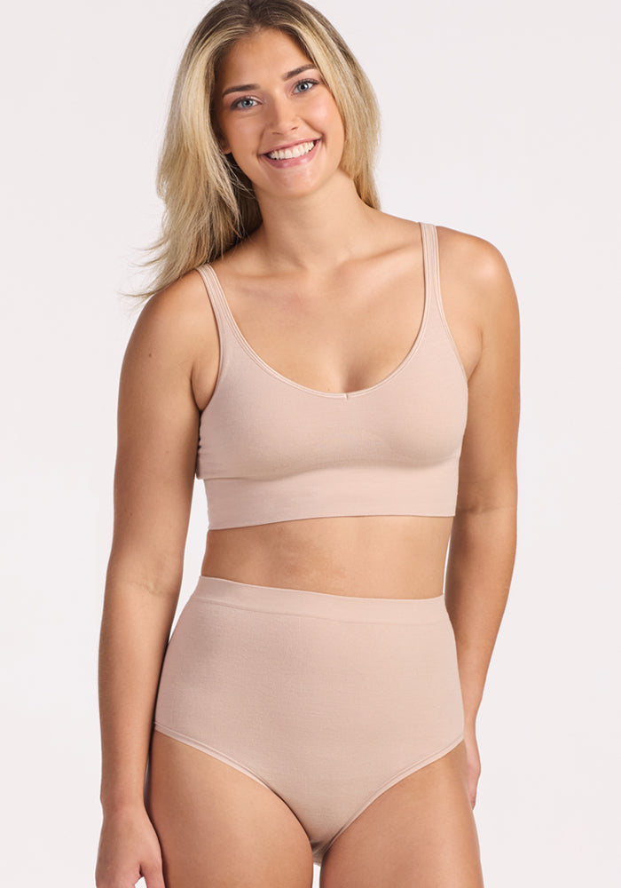A woman with long blonde hair smiles while wearing Woolx Mabel Full Coverage Underwear in Sand—a seamless nude-colored bralette and high-waisted undies—against a plain light background.