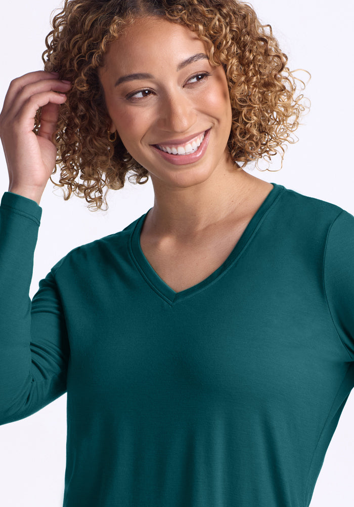 A smiling woman with curly hair wears the Woolx Millie Long Sleeve in Riverbank Green, touching her hair. The plain white background highlights the shirt’s temperature-regulating merino wool comfort.