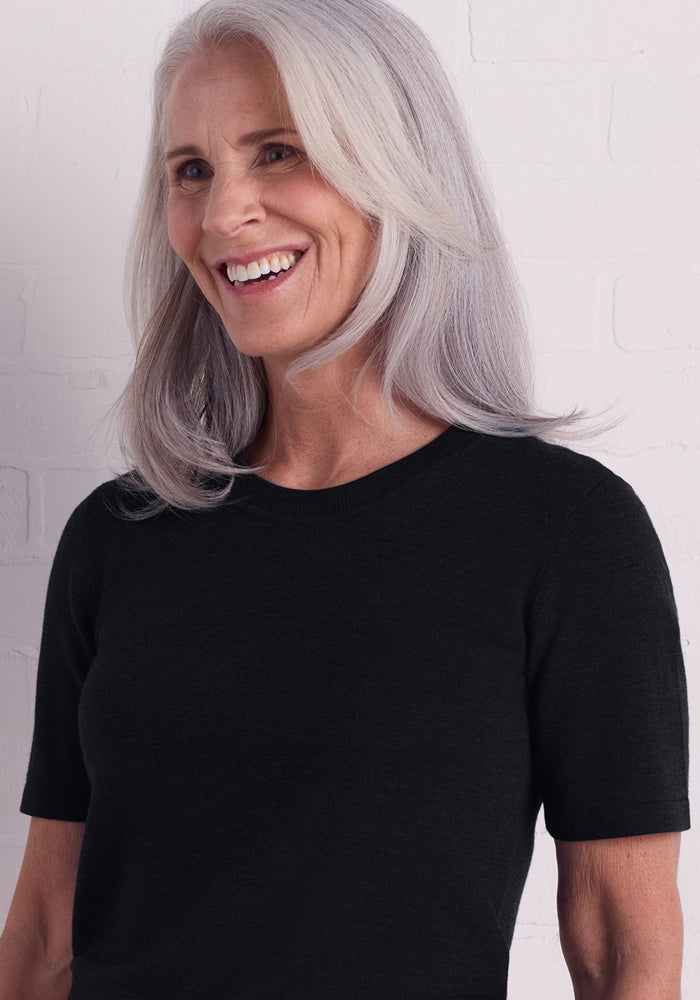 A smiling older woman with long, straight gray hair wears the Woolx Anna Crew Sweater in black as she stands before a white brick wall.